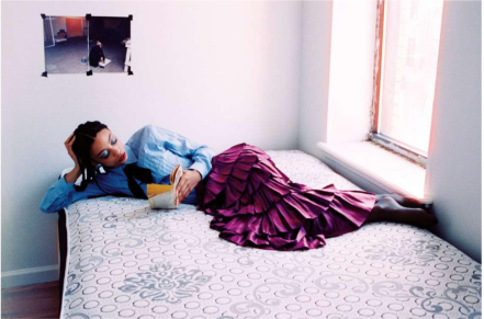 A woman in a ruffled, purple, ankle-length skirt and a blue blouse top reads while lying down on a bed in a sparsely decorated room filled with natural light from the bedside window.